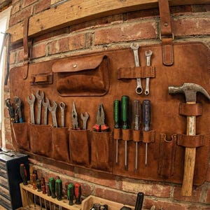 May include: A brown leather tool organiser with various tools, including spanners, screwdrivers, and a hammer, hanging on a brick wall. The organiser has multiple pockets and loops to hold the tools. The background shows a workshop setting.