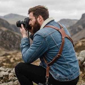 May include: A brown leather camera harness worn over a denim jacket. The harness has adjustable straps and metal hardware. The photographer is kneeling and holding a camera, with a mountainous landscape in the background. The harness is designed to carry a camera.