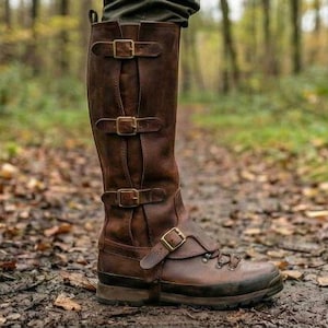 May include: Brown leather knee-high boot with multiple buckle straps. The boot has a rugged sole and is designed for outdoor use. The boot is shown on a forest path.