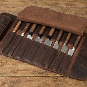 Op de afbeelding: Een bruine leren messenrol met een zakje met ritssluiting en verschillende messen met houten handvatten en zilveren lemmeten. De rol is open en toont de messen in afzonderlijke sleuven. De rol is vastgezet met leren riemen en gespen.