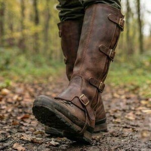 May include: Brown leather knee-high boots with multiple buckle closures. The boots have a rugged, outdoor design with a thick sole, suitable for walking on uneven terrain. The boots are worn on a muddy path in a forest setting.