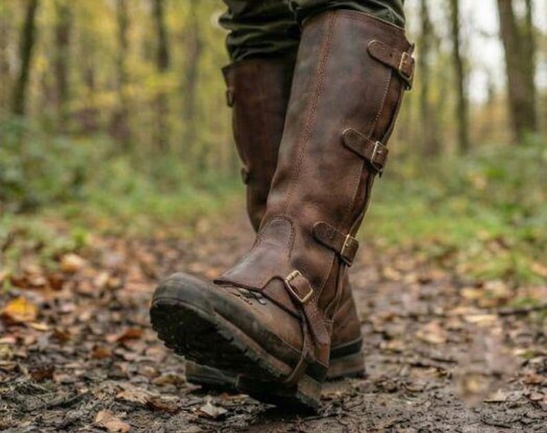 May include: Brown leather knee-high boots with multiple buckle closures. The boots have a rugged, outdoor design with a thick sole, suitable for walking on uneven terrain. The boots are worn on a muddy path in a forest setting.