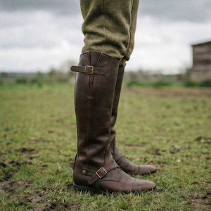 May include: Brown leather riding boots with buckle details. The tall boots are knee-high and have a classic design. The boots are worn with green tweed pants and are suitable for equestrian activities.