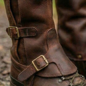 May include: Brown leather boots with brass buckles and laces. The boots have a rugged, worn appearance, with visible stitching and a textured surface. The design includes a strap and buckle detail around the ankle.