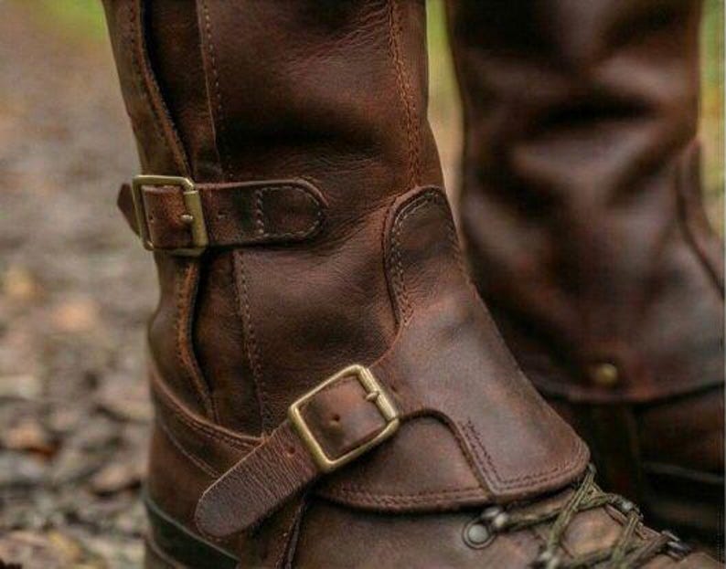 May include: Brown leather boots with brass buckles and laces. The boots have a rugged, worn appearance, with visible stitching and a textured surface. The design includes a strap and buckle detail around the ankle.
