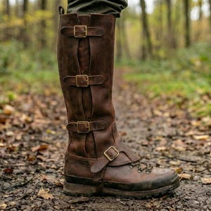 May include: Brown leather knee-high boots with multiple buckle closures. The boots have a rugged sole and are designed for outdoor use. The boots are shown on a forest path, suggesting they are suitable for hiking or similar activities.