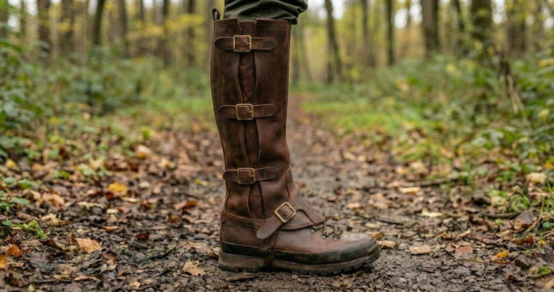 May include: Brown leather knee-high boots with multiple buckle closures. The boots have a rugged sole and are designed for outdoor use. The boots are shown on a forest path, suggesting they are suitable for hiking or similar activities.