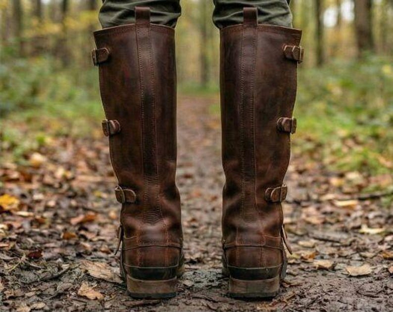 May include: A pair of brown leather knee-high boots with multiple buckle closures. The boots have a distressed finish and are worn on a path covered in fallen leaves. The boots are a classic style, suitable for outdoor activities.