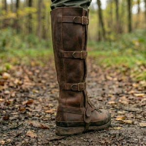 May include: A tall, brown leather boot with three buckle straps. The boot is worn on a muddy path in a forest setting. The boot appears to be durable and suitable for outdoor activities.