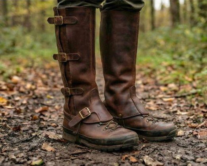 May include: A pair of brown leather knee-high boots with multiple buckle closures and a lace-up design on the foot. The boots are worn in a forest setting, with fallen leaves on the ground.