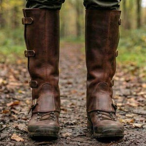 May include: A pair of brown leather knee-high boots with multiple buckle closures. The boots have a rugged, outdoor style with a lace-up design at the ankle. The boots are worn on a path covered in fallen leaves.