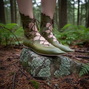 Peut inclure: Bottes en cuir vert avec des découpes en forme de feuilles et des lacets marron. Les bottes, à bout pointu, sont portées sur un rocher recouvert de mousse dans une forêt. Design de chaussures unique, inspiré de la nature.