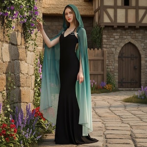 Puede incluir: Una mujer con un vestido negro de sirena y una capa con capucha. La capa tiene un degradado de verde y turquesa, con una capucha que enmarca la cara. El vestido es sin mangas y ajustado. El fondo muestra una pared de piedra y un edificio.