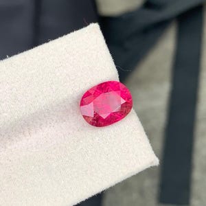 Könnte beinhalten: Ein leuchtender, oval geschliffener Edelstein mit einem tiefen, gesättigten pink-roten Farbton. Der Stein hat eine facettierte Oberfläche, die das Licht reflektiert und ein brillantes, funkelndes Aussehen erzeugt. Er liegt auf einem hellen, strukturierten Stoff.