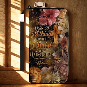 Puede incluir: Una cartera negra con correa de muñeca con un diseño floral y de león. La cartera tiene el texto "I can do all things through Christ who strengthens me" y "Philippians 4:13".