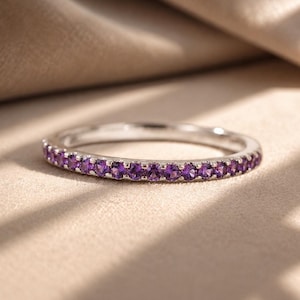 Op de afbeelding: Een dunne zilveren ring met een doorlopende rij kleine, rondgeslepen paarse amethist edelstenen die langs de voorste helft van de metalen band zijn gezet. De ring heeft een gepolijste metalen afwerking en een slank profiel.