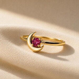 Könnte beinhalten: Ein goldener Ring mit einem Mondsichel-Design und einem einzelnen runden, facettierten, leuchtend pinkfarbenen Edelstein, der in die Kurve des Mondes eingefasst ist. Die Ringschiene ist dünn und poliert und weist ein glattes, reflektierendes Finish auf.