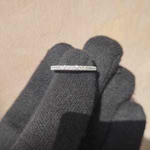 May include: A thin, silver ring with a row of small, clear gemstones. The ring is held by a gloved hand, showcasing the delicate design. The ring appears to be a wedding band or a similar type of jewelry.