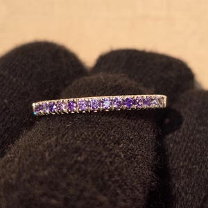 Könnte beinhalten: Ein silberner Ring mit einer Reihe kleiner, violetter Edelsteine. Der Ring wird vor einem dunklen, strukturierten Hintergrund gehalten, der das Funkeln der Edelsteine hervorhebt. Der Ring ist ein Schmuckstück.