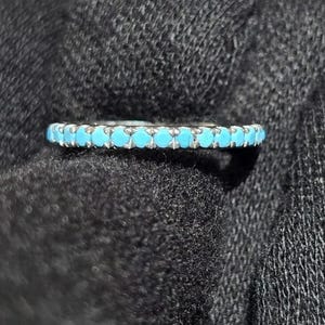 Op de afbeelding: Een zilveren ring met een band van kleine, ronde, heldere turkooizen stenen. De ring is geplaatst tegen een donkergrijze, getextureerde achtergrond.