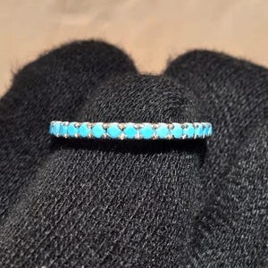 May include: A silver ring with a band of small, bright turquoise stones. The ring is set against a dark, textured background, highlighting the vibrant blue of the gemstones. The ring is a piece of jewelry.