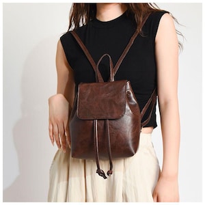 Può includere: Uno zaino in pelle marrone con chiusura a coulisse e patta. Lo zaino ha spallacci regolabili e una maniglia superiore. La pelle ha un aspetto strutturato. La borsa è indossata sulle spalle.