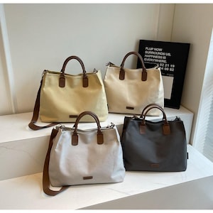 Può includere: Quattro borse tote in vari colori: beige, bianco sporco, grigio chiaro e grigio scuro. Ogni borsa ha manici marroni e una tracolla abbinata. Un cartello nero con testo bianco è sullo sfondo.