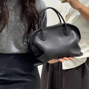 May include: A black handbag with a smooth, rounded silhouette and two top handles. The bag features a zippered closure along the top edge, a soft structure, and a minimalist design with clean lines and no visible external pockets or hardware.