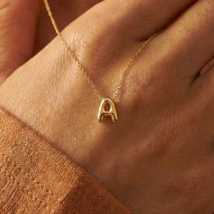 Op de afbeelding: Een delicate gouden ketting met een hanger in de vorm van de letter "A". De hanger is een afgeronde hoofdletter "A" die aan een fijne gouden ketting hangt. De ketting wordt getoond tegen een warme, neutrale achtergrond.