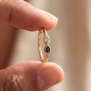 May include: A gold ring featuring two teardrop-shaped gemstones, one light blue and one dark blue, set in an infinity symbol design. The ring is held between fingers, showcasing its delicate details and elegant style.