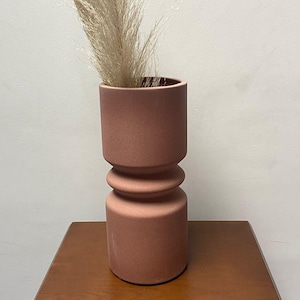 May include: A terracotta-colored ceramic vase with a cylindrical top and base, and a rounded midsection. The vase holds dried, beige-colored ornamental grass. The vase sits on a wooden table.