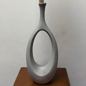 Op de afbeelding: Een grijze keramische vaas met een ovale uitsparing en een smalle hals, met gedroogd siergras. De vaas staat op een houten oppervlak tegen een effen witte muur. De vaas is ongeveer 30 cm hoog.