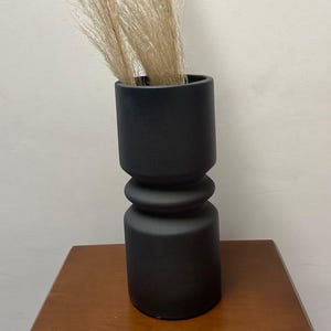 Può includere: Un vaso nero moderno con forma cilindrica e design impilato. Il vaso contiene diversi steli di erba della pampa beige essiccata. Il vaso è appoggiato su una superficie di legno.