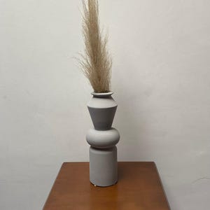 Op de afbeelding: Een grijze, geometrische vaas met een smalle hals en een brede basis, met gedroogd pampasgras. De vaas staat op een donkerbruin houten nachtkastje met twee lades. De achtergrond is een effen witte muur.