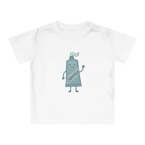 Op de afbeelding: Een wit t-shirt met korte mouwen en een centrale afbeelding van een geanimeerde tandpastatube. De blauwgrijze tube heeft een lachend gezicht, dunne armen en benen, en het woord SQUEASY staat diagonaal in donkerblauw op de voorkant.