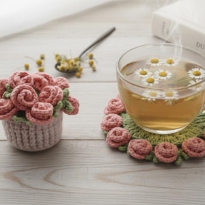 Pode incluir: Uma chávena de chá de vidro transparente cheia de chá de camomila e flores flutuantes, colocada sobre um descanso de copo de crochê com rosas cor-de-rosa. Uma cesta de flores de crochê com rosas cor-de-rosa está ao lado. Uma colher com flores de camomila está no fundo.