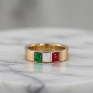 Puede incluir: Un anillo de banda en tono dorado con tres piedras incrustadas de corte cuadrado dispuestas horizontalmente. Las piedras son de color verde, blanco y rojo, creando un patrón tricolor en la parte frontal del anillo de metal liso y pulido.