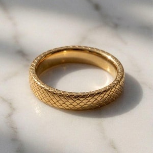 May include: A gold-toned ring featuring a textured, cross-hatched pattern across its outer surface. The band has a consistent width and a polished finish, reflecting light across the repeating diamond-shaped grid design on the metal exterior.
