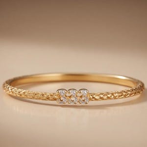 Peut inclure: Un bracelet jonc doré avec une surface texturée et un motif central de trois figures connectées, chacune ornée de petites pierres précieuses claires. Le bracelet est posé sur un fond doux et neutre, soulignant son design élégant.