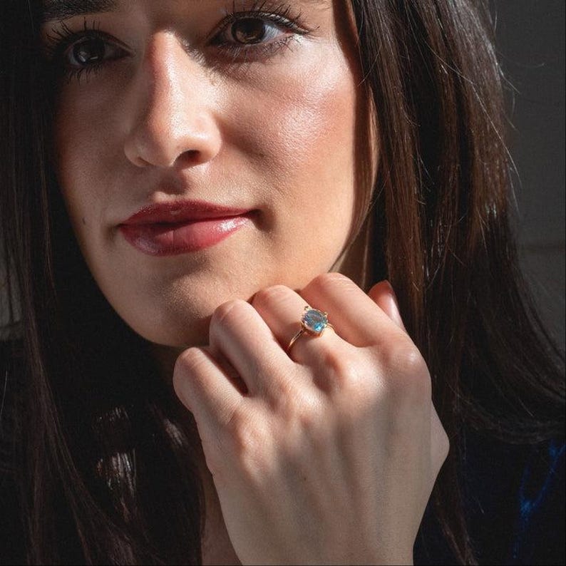 May include: A gold ring with a light blue gemstone. The ring is on a person's finger. The gemstone is oval-shaped and set in a gold band. The person has long brown hair and is wearing makeup.