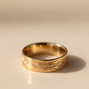 Könnte beinhalten: Ein goldfarbener Ring mit einem eingravierten Sonnenstrahlen- und Wellenmuster. Der Ring besteht aus einem glänzenden, reflektierenden Metall und liegt auf einer neutral getönten Oberfläche. Das Design des Rings weist ein sich wiederholendes Muster um das Band auf.