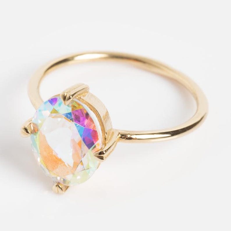 May include: A gold-colored ring featuring a large, iridescent, oval-cut gemstone. The gemstone displays a spectrum of colors, including pink, blue, and yellow. The ring has a simple, thin band and a four-prong setting.