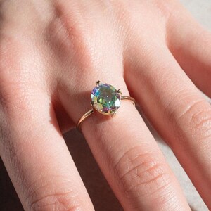 May include: A gold ring featuring an oval-cut, iridescent gemstone. The gemstone displays a spectrum of colors, including green, blue, and purple. The ring's band is a thin, polished gold. The ring is shown on a finger.