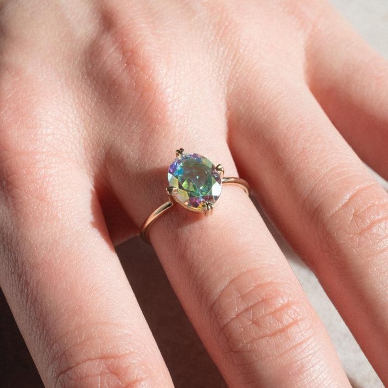 May include: A gold ring featuring an oval-cut, iridescent gemstone. The gemstone displays a spectrum of colors, including green, blue, and purple. The ring's band is a thin, polished gold. The ring is shown on a finger.