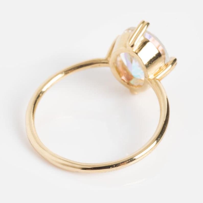 May include: A gold ring with a large, iridescent gemstone. The gemstone has a round cut and displays a range of colors. The ring's band is thin and polished, and the setting has four prongs.