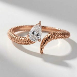 May include: A rose gold bypass ring featuring a textured, scale-like pattern along the band. One end of the open band terminates in a pear-shaped, clear faceted gemstone, while the other end tapers to a point, mimicking the appearance of a snake.