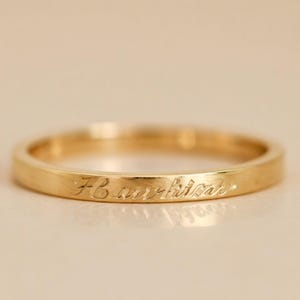 Könnte beinhalten: Ein goldener Bandring mit graviertem Text. Der Ring hat ein schlichtes, elegantes Design und eine polierte Oberfläche. Der Text scheint ein Name oder Initialen zu sein. Der Ring ist wahrscheinlich aus Gold.