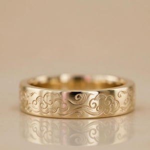 Puede incluir: Un anillo dorado con un diseño de nubes grabado. El anillo tiene un acabado pulido y una banda ancha. El diseño de nubes está grabado alrededor de todo el anillo.