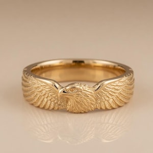 May include: A gold-colored ring featuring a detailed eagle design. The eagle's head and wings are prominently displayed, with intricate feather detailing. The ring has a polished finish and a band with a textured pattern.