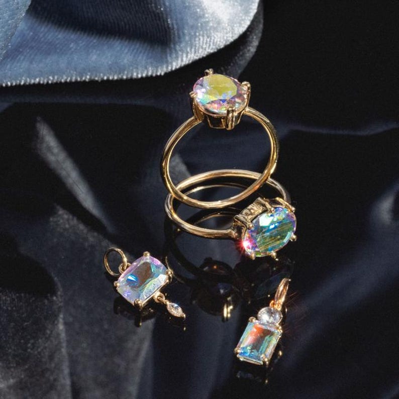 May include: Gold rings and pendants with iridescent, oval and rectangular gemstones. The rings are stacked, and the pendants have small, dangling accents. The jewelry is displayed on a black surface with a blue velvet backdrop.